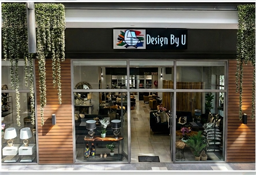 The DesignByU shopfront at Design Quarter in Fourways, its lit window display showing a dining table, upholstered chairs and pendant lighting behind the glass.